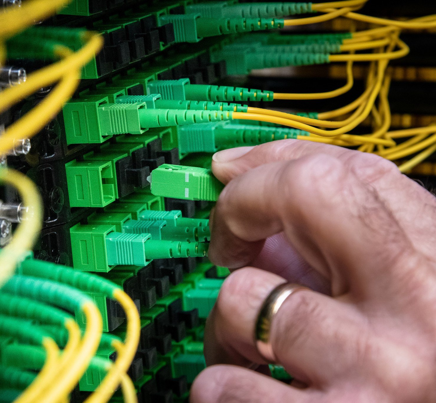 A technician's hand plugging a green fibre-optic cable into a network cabinet full of patch cables