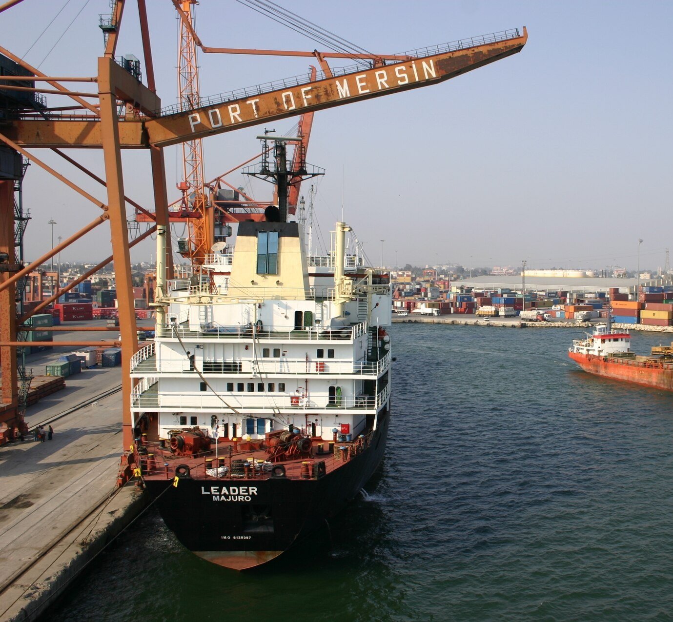 A cargo ship alongside the quay beneath the cranes of the Port of Mersin, Türkiye, with stacked containers behind it